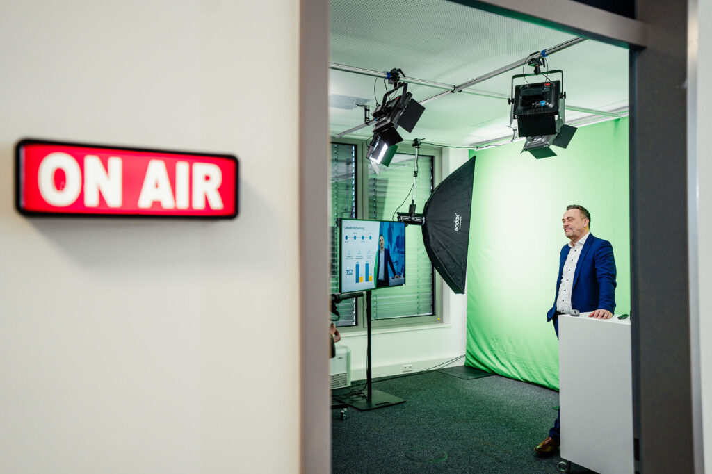 Thomas Grimm in kleinem Videostudio vor Greenscreen, Türschild "On Air".