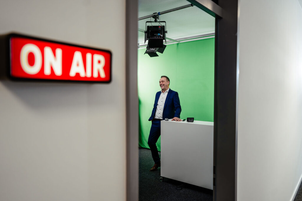 Thomas Grimm in kleinem Videostudio vor Greenscreen, Türschild "On Air".